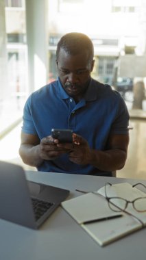 Ofisteki genç Afro-Amerikalı adam telefonuna dizüstü bilgisayar ve not defteriyle bakıyor.