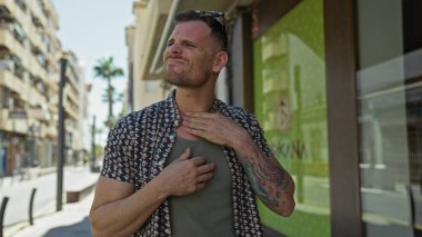 Dövmeli yakışıklı sakallı adam güneşli şehir caddesinde boğaz ağrısı çekiyor.