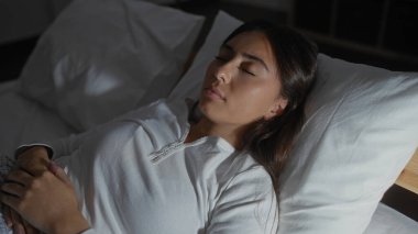 Genç bir kadın, esmer saçlarıyla yatakta huzur içinde uyuyor. Yatak odasında beyaz bir bluz giyiyor. Kapalı alanda sakin bir rahatlama sahnesi sergiliyor.