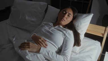 Genç İspanyol kadın yatakta uzanıyor. Yatak odasının iç kısmında, esmer saçlı ve beyaz pijamalı kötü görünüyor..