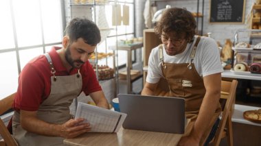 Pastanenin içinde birlikte çalışan adamlar önlük takan iki fırıncı belgeleri inceliyor ve dükkanın içinde dizüstü bilgisayar kullanıyorlar.