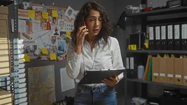 A concerned middle-aged woman detective analyzes evidence in a cluttered investigation office, talking on the phone.