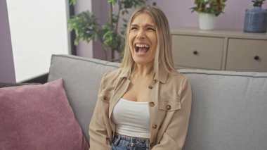 Genç, güzel, sarışın bir kadın yukarı bakarken çığlık atıyor, modern bir oturma odasında, içinde çiçekler ve mor bir yastıkla oturuyor, ev dekorasyonunu sergiliyor..