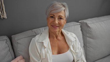 Oturma odasındaki kanepede oturan, gülümseyen ve kameraya bakan, güzel, olgun ve rahat bir yüz ifadesini sergileyen, kısa gri saçlı yaşlı kadın..