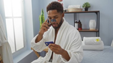 Burun bandı ve bornoz takan genç bir adam, telefonla konuşuyor ve bir kaplıca odasında kredi kartı tutuyor..