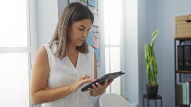 Ofis odasında tablet kullanan genç bir kadın, modern bir iş ortamında kapalı yeşil ve beyaz duvarlı odaklanmış bir ifade gösteriyor.