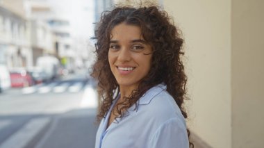 Şehir sokağında poz veren genç bir kadın güzel, kıvırcık esmer saçlarını şehir zemininde sergiliyor.