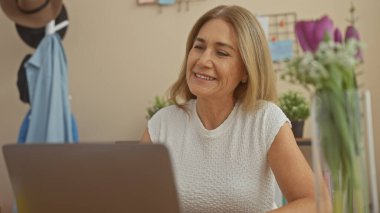 Modern bir ofis ortamında dizüstü bilgisayarla çalışan gülümseyen orta yaşlı beyaz bir kadın.