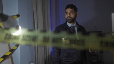 Ciddi bir İspanyol sakallı adam, loş bir oturma odasında sarı polis şeritleriyle sarılı bir suç mahallini araştırıyor..