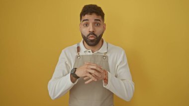 Sarı arka planda duran önlük takan sakallı yakışıklı İspanyol adam şaşırmış görünüyor.