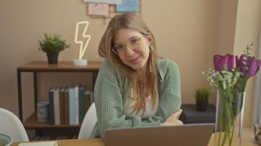 Gülümseyen, gözlüklü, dizüstü bilgisayarlı, etrafı dekoratif elementler ve mor çiçeklerle çevrili genç bir kadının samimi bir portresi..