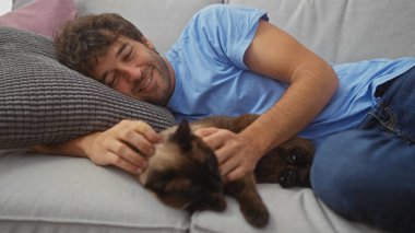 İspanyol bir adam rahat bir ortamda kanepede siyam kedisiyle sarılıp yatıyor..