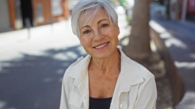 Güzel gri saçlı kadın açık havada gülümsüyor açık bir şehir sokağında, neşeli ve olgun bir varlığı canlandırıyor şehir ortamında.