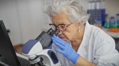 Senior woman scientist examining specimen through microscope in laboratory setting. clipart