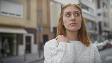 Düşünceli, kızıl saçlı, süveter giyen genç bir kadın şehir sokaklarında poz veriyor..
