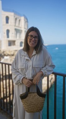 Polignano 'da sepetle poz veren genç, güzel bir İspanyol kadın çarpıcı bir mavi denize ve tarihi mimariye karşı kısrak, puglia, italyan.