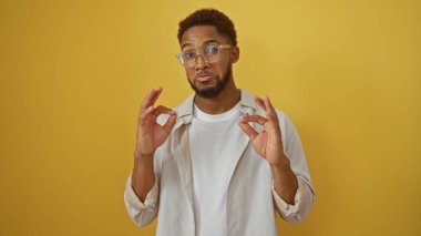 Genç adam, gözlüklü ve günlük kıyafetlerle izole edilmiş sarı arka planda iyi bir jest yapıyor.