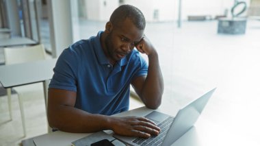 Ofis odasında laptopla çalışan genç Afro-Amerikan bir adam kapalı alanda odaklanmış görünüyor.