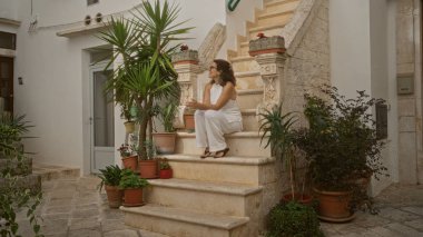 Taş basamaklarda oturan genç ve güzel bir İspanyol kadın büyüleyici eski Locorotondo, Puglia, İtalya 'da bitkilerle çevrili Avrupa sokak hayatının özünü yakalıyor..