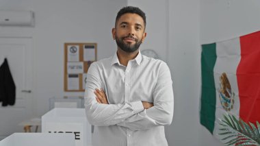 Sakallı, kendine güvenen genç adam kollarını kapalı bir yerde beyaz bir gömlek içinde ve arka planda Meksika bayrağı ile çapraz duruyor..