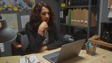 Ofisinde dizüstü bilgisayarı olan düşünceli, orta yaşlı bir İspanyol kadın. Etrafı dedektif soruşturması ipuçlarıyla çevrili..