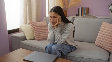 Oturma odasında kanepede oturan bir kadın masadaki dizüstü bilgisayara endişeli bir şekilde bakıyor. Arka planda yastıklar ve kitaplıklar var.