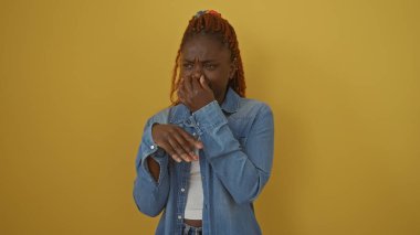 Kot ceketli genç siyah bir kadın burnunu çimdikliyor ve sarı bir arka plana iğrenç bir ifade takınıyor..