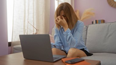 Oturma odasında oturan genç bir kadın kendini stresli hissederken dizüstü bilgisayarı üzerinde çalışırken iç dekorasyon ve dekorasyon unsurlarıyla çevrili bir şekilde oturuyor..