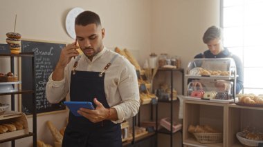 Bir pastanede çalışan erkekler, biri telefon alıyor ve elektronik tablet yönetiyor diğeri ise modern, iyi aydınlatılmış bir iç mekanda hamur işleri düzenliyor.