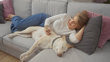 Genç bir kadın, köpeği ile birlikte rahat bir ev ortamında, akıllı bir telefon tutarak kanepede dinleniyor..