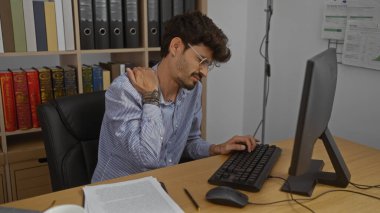 Genç, sakallı, boynu ağrıyan bir adam. Ofis ortamında bir bilgisayarda çalışırken. Etrafı kitaplık ve evrak işleriyle çevrili.