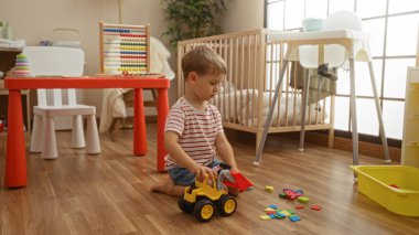 Beyaz çocuk oyuncak kamyonla çeşitli eğitim oyuncakları ve mobilyalarla dolu bir anaokulu sınıfının zemininde oynuyor.