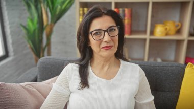 Gözlüklü, olgun bir İspanyol kadın, rahat bir oturma odasındaki koltukta, düşünceli bir şekilde oturur..