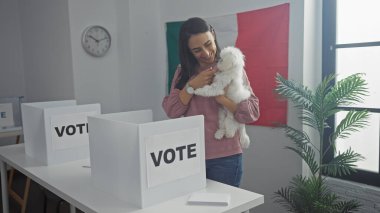 Meksikalı bir oy kullanma merkezinde elinde bichon köpeğiyle gezen İspanyol bir kadın kapalı mekanlarda ve bayrağıyla gülümsüyor..