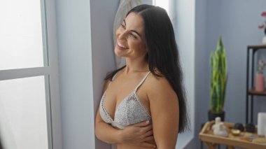 Pencerenin yanında beyaz iç çamaşırı giymiş, gülümseyen bir kadın.