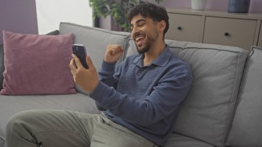 Evdeki koltukta akıllı telefon kullanan neşeli genç adam heyecanını dile getiriyor..