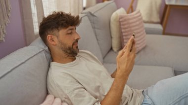 Kafkasyalı genç bir adam akıllı telefon kullanırken bir kanepede dinleniyor. Yastıklarla ve doğal ışıkla evin içini tasvir ediyor..