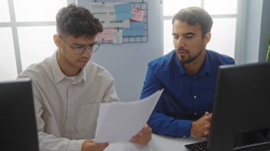 Ofiste birlikte çalışan iki adam bilgisayarlı ve arka planda bir strateji kurulu olan belgeleri analiz ediyor.