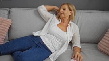 Rahat yastıklı, rahat bir oturma odasında kanepede dinlenen, rahat kıyafetler giyen ve evde huzurlu bir anın tadını çıkaran bir kadın..