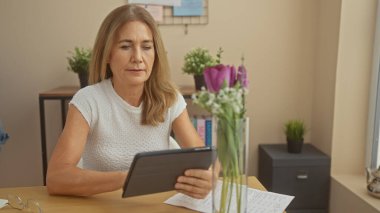 Olgun bir kadın, rahat bir oturma odasında, masada çiçekler, gözlükler ve evraklarla kitap okur..