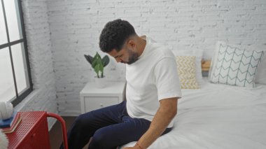 Genç adam modern yatak odasında beyaz tuğla duvar ve desenli yastıklarla oturuyor.