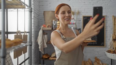 Fırında selfie çeken bir kadın elinde ekmek, önlük, etrafını somunlar, raflar ve ev ortamında karatahta ile çevrili.