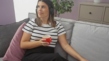 Genç bir kadın evinde rahat bir oturma odasında oturup kırmızı bardaktan içiyor ve kulaklıkla müzik dinliyor..