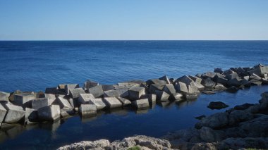 Ocean view from the rocky shore of gallipoli, puglia, italy with concrete tetrapods lining the coast under a clear blue sky. clipart