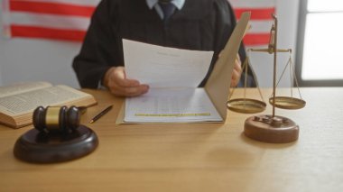 Male judge in courtroom reviewing documents with gavel and scales of justice on desk before american flag clipart