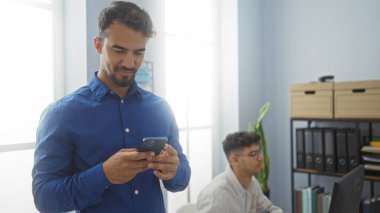 Bir iş ofisinde çalışan İspanyol adamlar, biri telefon kullanıyor ve diğeri bilgisayar üzerinde çalışıyor.