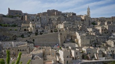 Materya, Basilicata, İtalya 'daki antik yamaç manzarasında tarihi taş binalar ve açık mavi gökyüzünün altında bir kilise kulesi var..