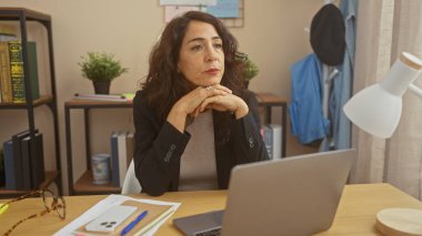 Düşünceli, orta yaşlı, İspanyol bir kadın ev ofisinden bilgisayarı, gözlüğü ve günlük kıyafetleriyle çalışıyor..