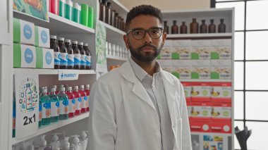 Genç, sakallı, beyaz önlüklü Afro-Amerikan bir adam modern bir eczane içinde güvenle duruyor.