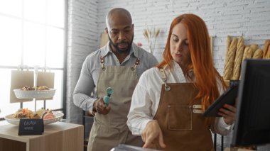 Erkek ve kadın birlikte bir fırında çalışıyor, hamur işleri düzenliyor ve sergilenen tuğla duvarlar ve taze ekmekle birlikte kapalı bir dükkanda envanter işlemek için bir tablet kullanıyor.